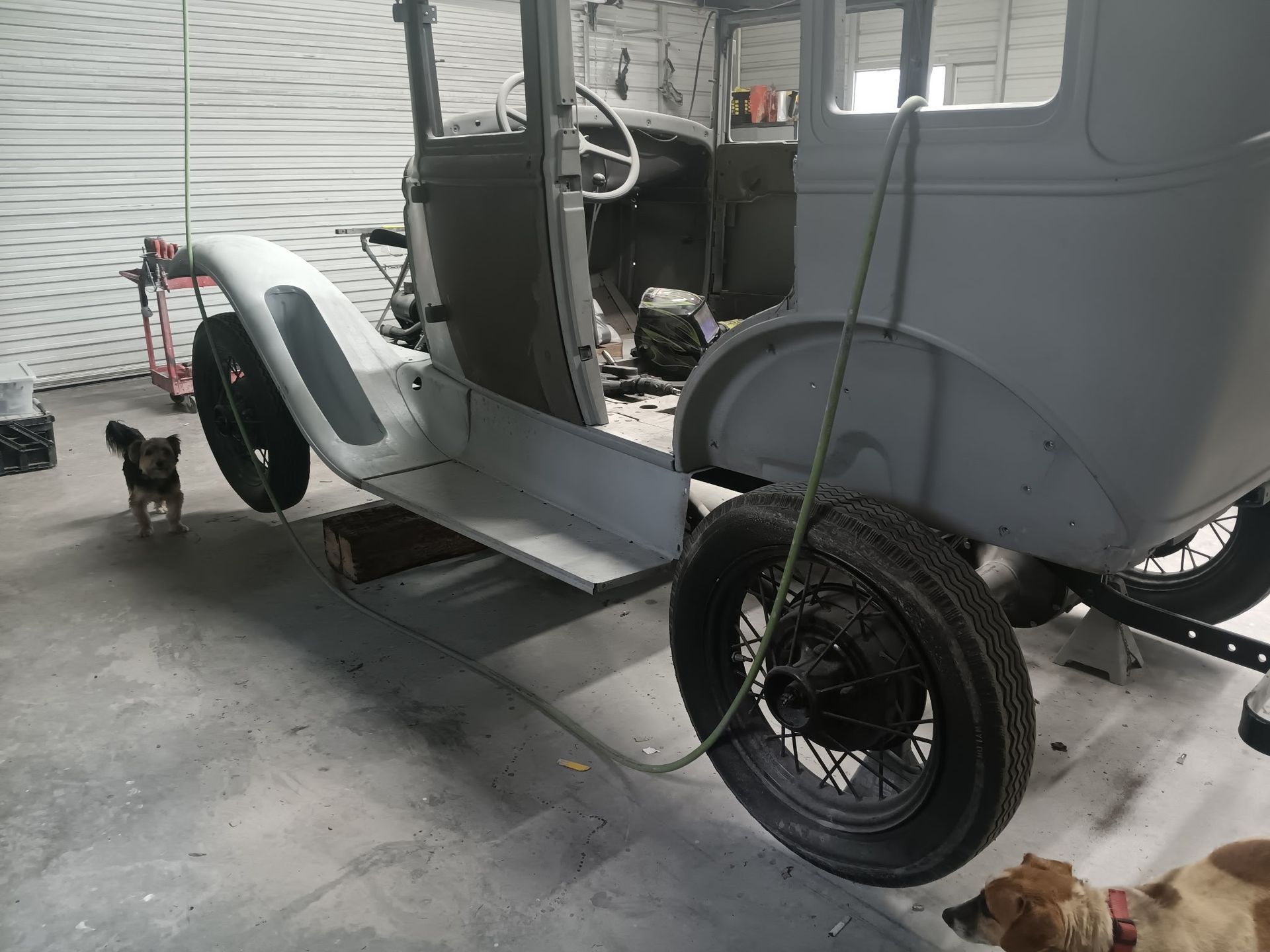 A gray vintage car frame is being restored in a garage with two dogs nearby.