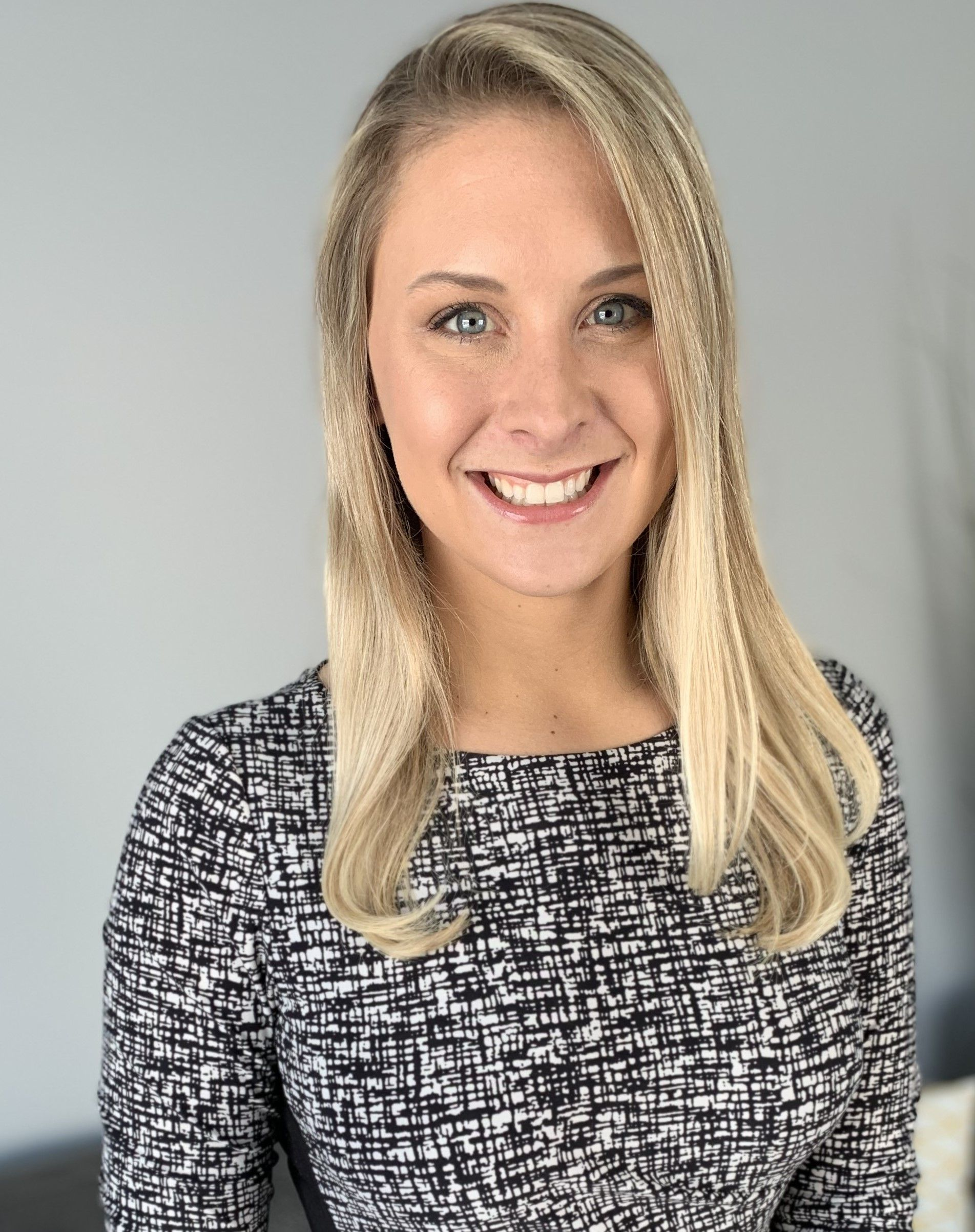 Woman with blonde hair smiling, wearing a black and white patterned top, standing in front of a gray wall.