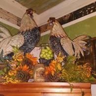 Two decorative roosters on a floral arrangement above a wooden mantel