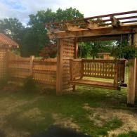 Wooden pergola and porch swing in a backyard with a fence and grassy yard.