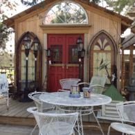 Rustic wooden cabin with red doors and arched windows, viewed from a deck with white outdoor chairs and table