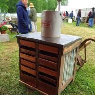 Outdoor wooden cart with a metal milk can on top at a grassy fairground.