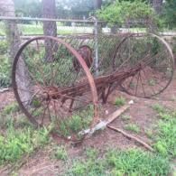 Rusty old farm equipment with large wheels overgrown with grass in a muddy yard