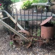Rusty old garden tiller with wooden handles beside a black iron fence in a yard.