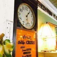 Wall clock above a vintage orange-crush sign beside a lit table lamp in a cozy interior.