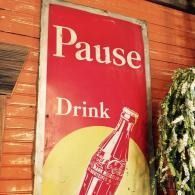 Red poster reading “Pause Drink” with a soda bottle and sunburst graphic on a wooden wall