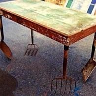 Old wooden farm table with rusted tools hanging beneath it on a pavement.