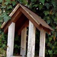 Wooden birdhouse with a peaked roof, weathered white posts, and ivy in the background