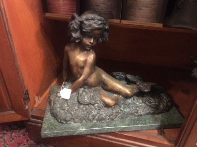 Small bronze child statue seated on a green base beside books on a wooden shelf.