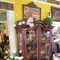 Decorative wooden cabinet with glass doors, topped with pink flowers, in a warmly lit antique shop
