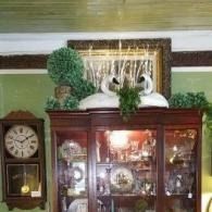 Wooden china cabinet with glass doors, topped by white ceramic swans, greenery, and a topiary beside a wall clock.