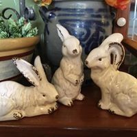 Three white ceramic rabbit figurines on a wooden shelf, with a blue vase and potted plant behind them