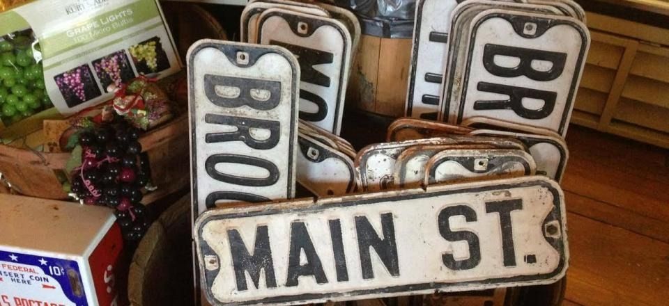 Stacked vintage street signs reading “MAIN ST.” and “BRO...” in a shop display