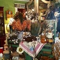 Cluttered shop display with colorful flowers, pottery, and decorative items in a cozy indoor setting