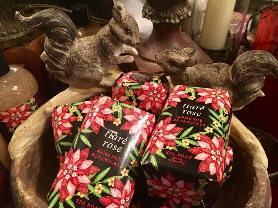 Assorted rose-scented soap bars in floral packaging in a decorative bowl, with candles and a squirrel figurine nearby