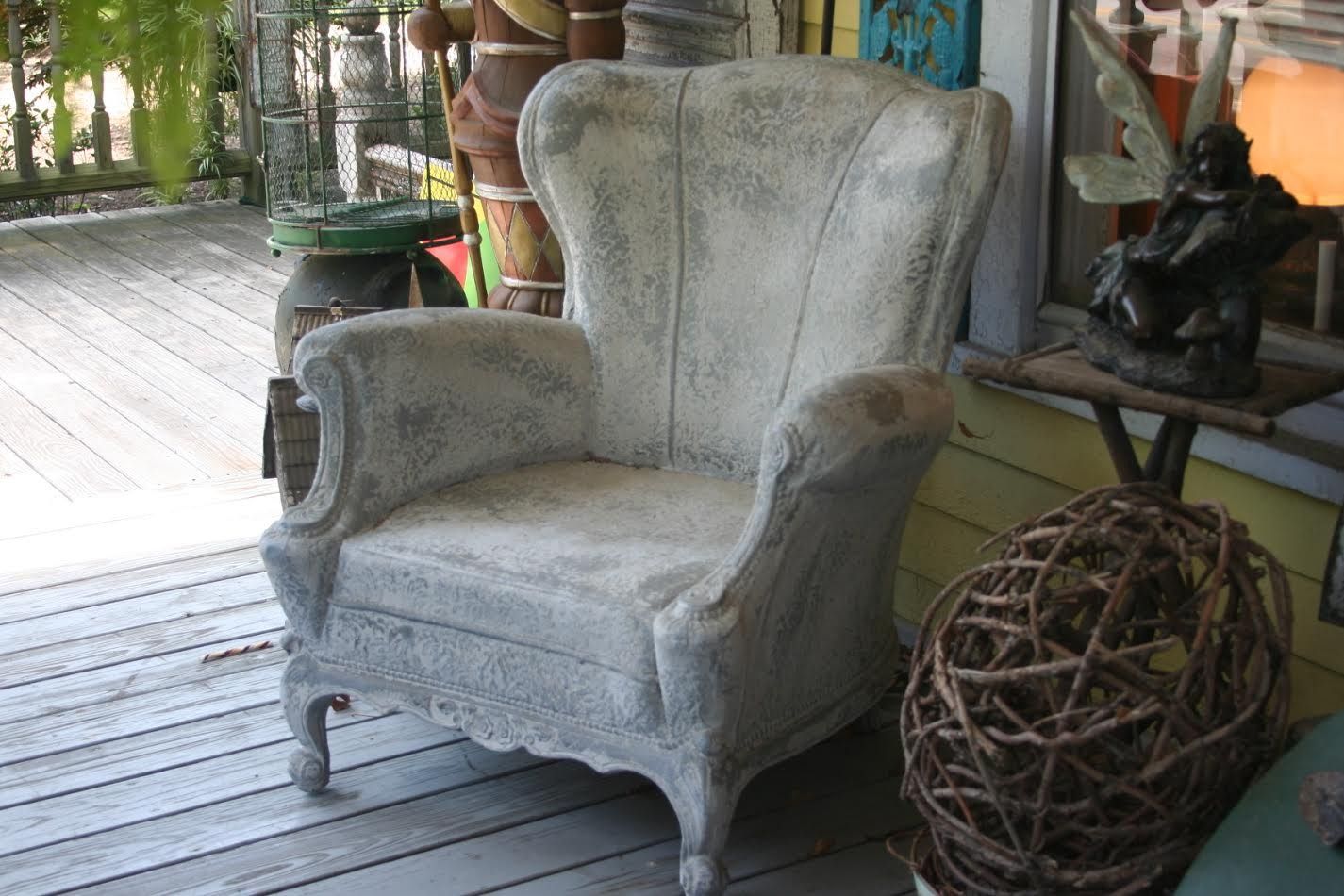 Gray upholstered wingback chair on a wooden porch beside a round wicker basket and other rustic decor.
