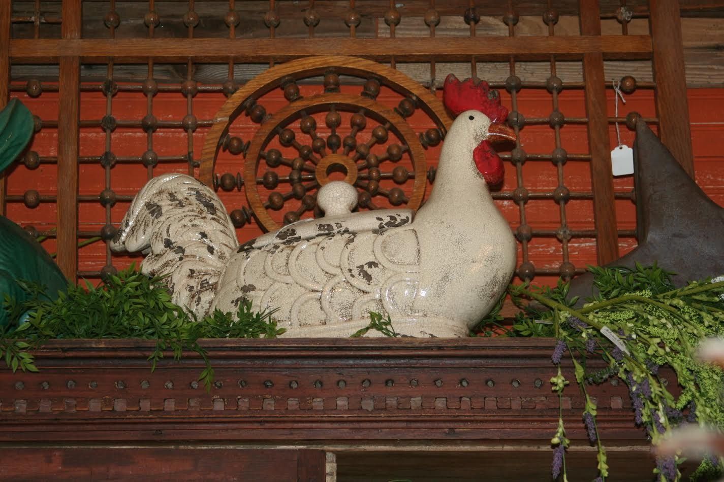Decorative white rooster statue with red comb on a wooden shelf, framed by greenery and a lattice backdrop