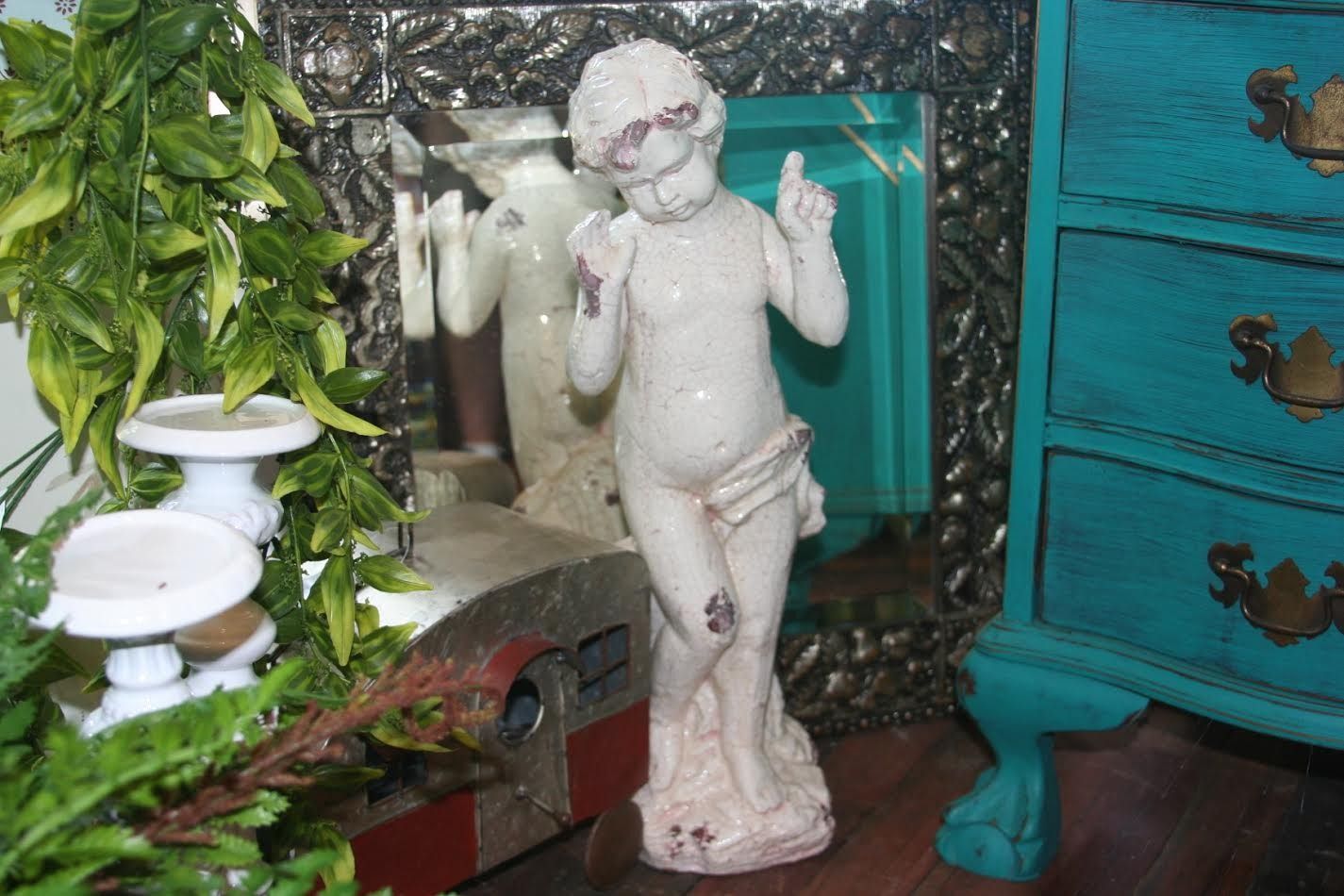 White cherub figurine beside leafy decor and teal dresser drawers in a miniature room scene