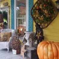 Porch with autumn decor, including a large pumpkin, wreath, and vintage furniture by a yellow house