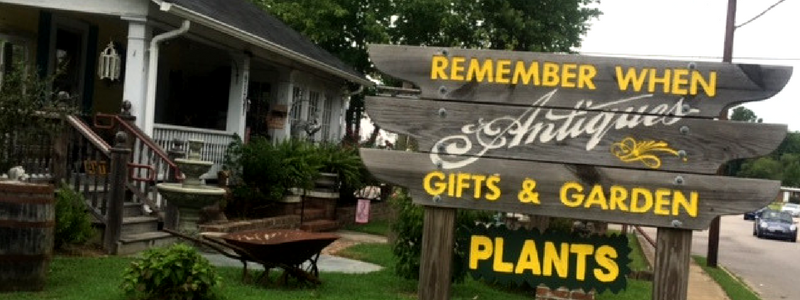 Rustic garden shop sign reading “Remember When Antiques, Gifts & Garden Plants” beside a porch