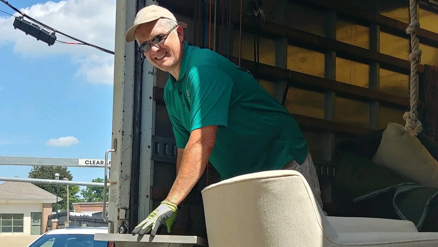 Man in green shirt and cap loading a rolled-up rug into a truck, smiling outdoors.