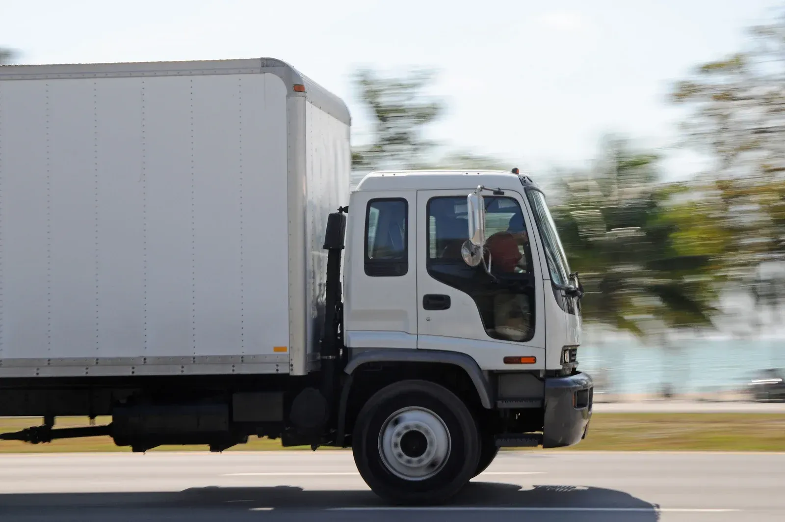 White box truck driving on a road; driver visible inside; trees in the blurred background.