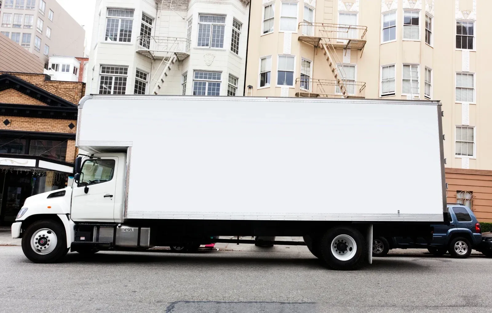 White moving truck parked on a city street, with apartment buildings in the background.