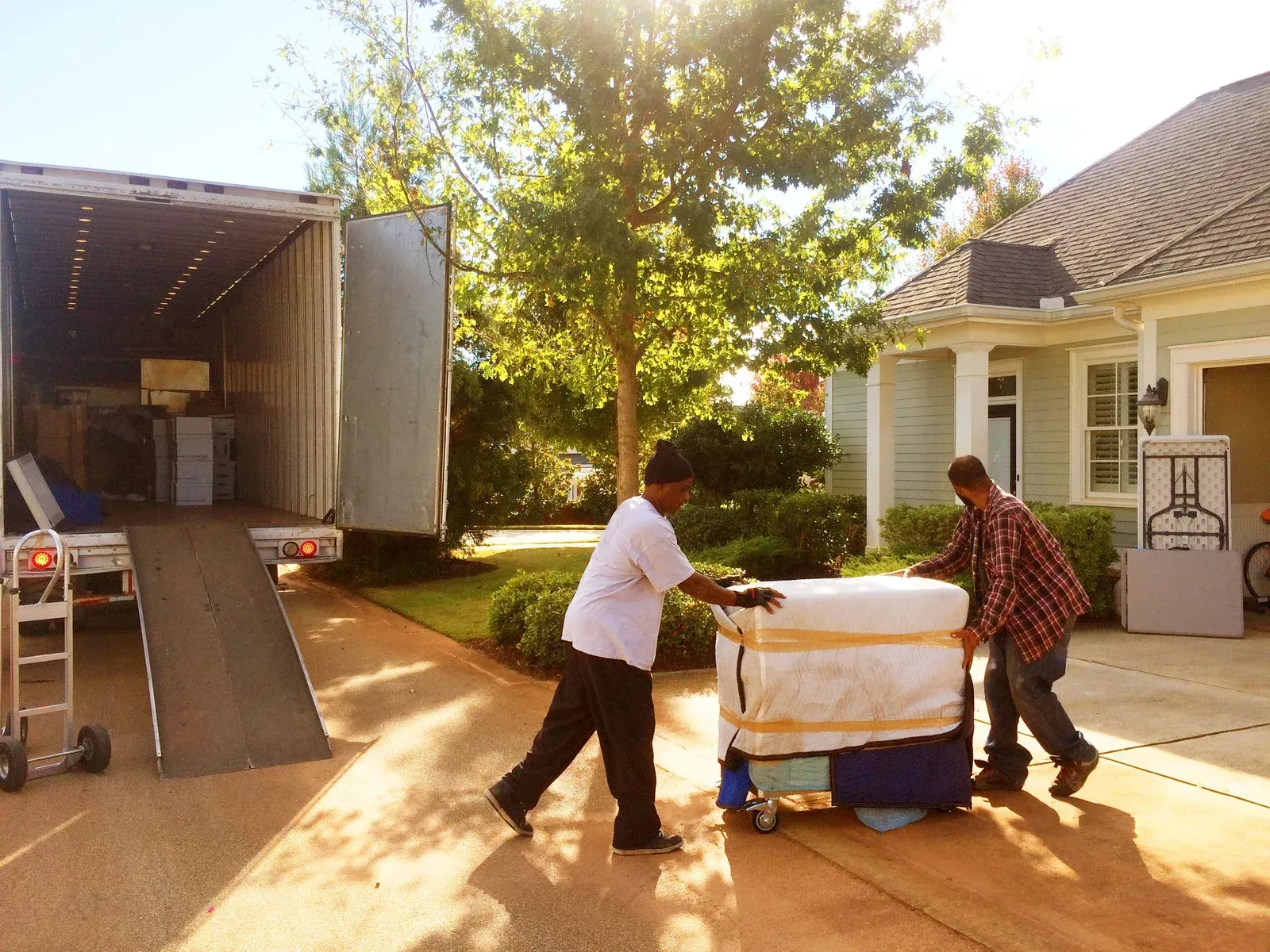 Two men are pushing a refrigerator on a dolly in front of a house.