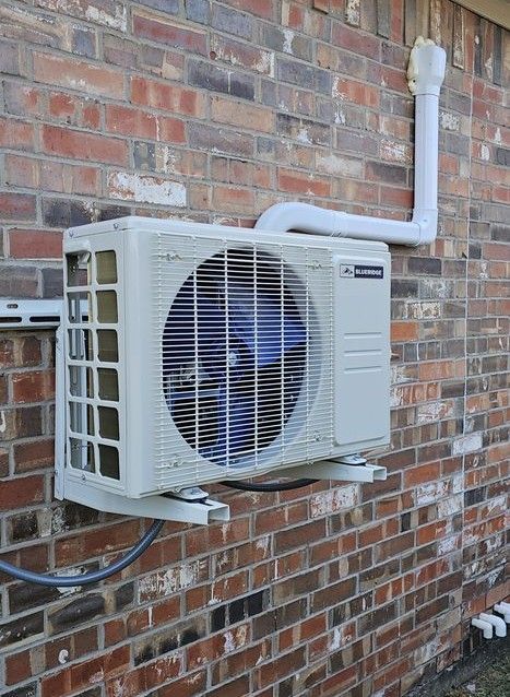 Outdoor air conditioning unit attached to a brick wall, with a white exhaust pipe.