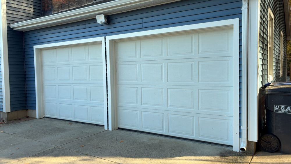 A house exterior with two white panelled garage doors on a blue-sided building next to a trash bin.