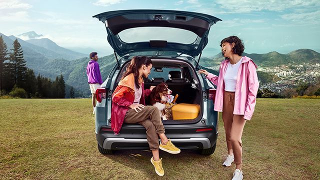 Pessoas com um cachorro reunidas ao redor do porta-malas aberto de um carro, ao ar livre, com uma montanha ao fundo.