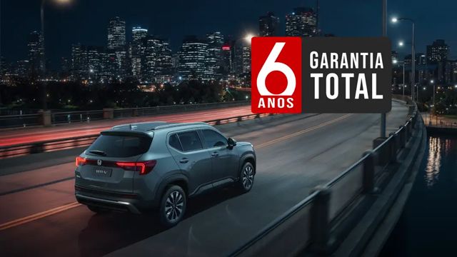 SUV cinza atravessando uma ponte à noite; anúncio de garantia total de 6 anos. Horizonte da cidade ao fundo.