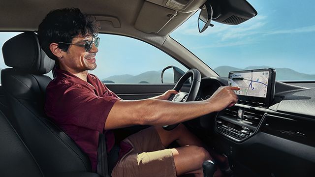 Homem de óculos escuros dentro de um carro, sorrindo para a tela de navegação, em um ambiente externo ensolarado.