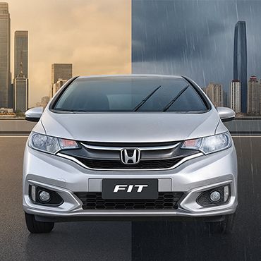 Carro Honda Fit prateado em paisagem urbana, com climas contrastantes: ensolarado à esquerda, chuvoso à direita.