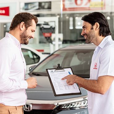 Homem de camisa branca analisa documento com mecânico em um centro de serviços Honda, apontando para o formulário.