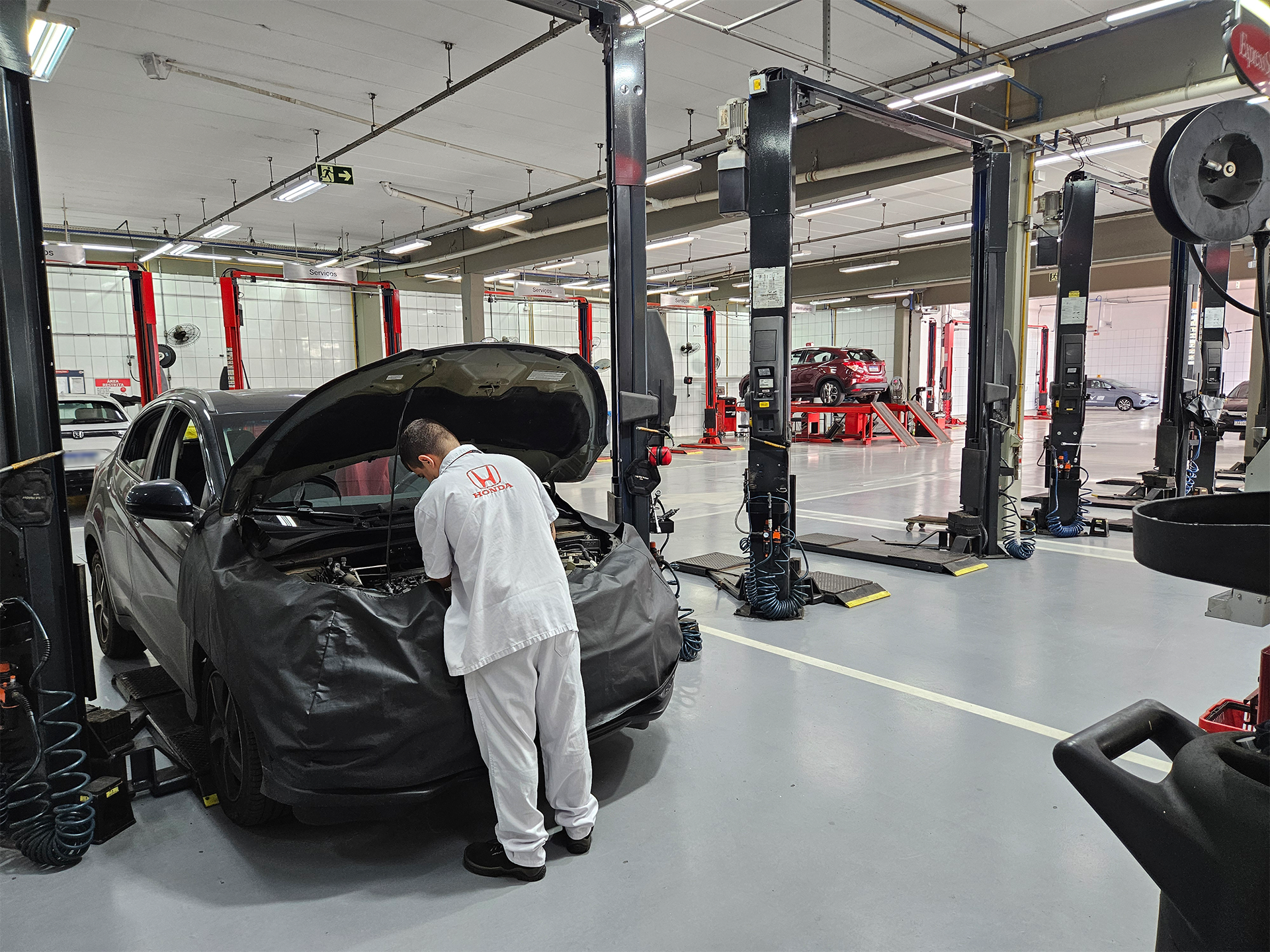 Mecânico trabalhando em um carro em uma oficina, com outros carros e equipamentos visíveis.