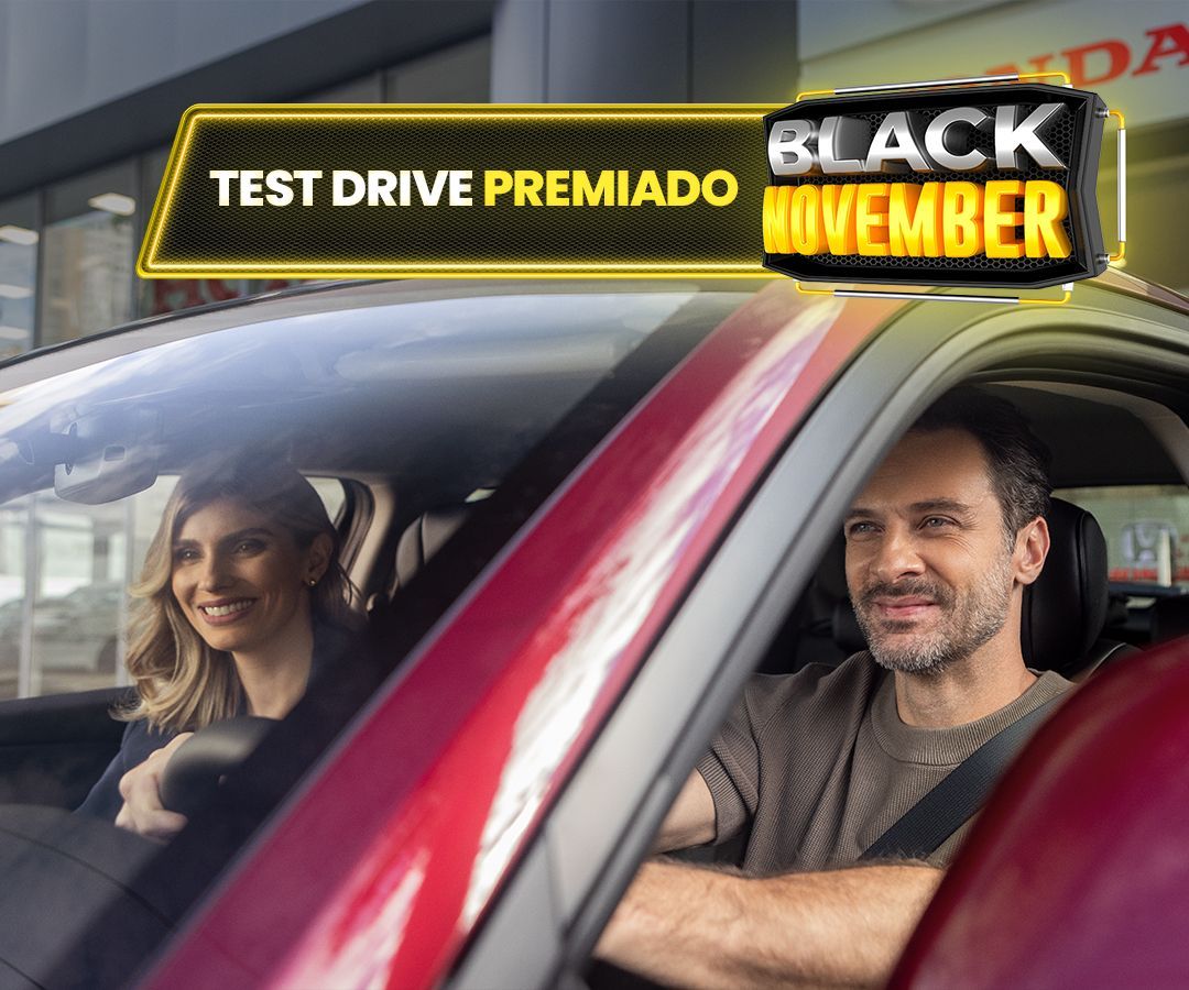 Homem de óculos escuros dentro de um carro, sorrindo para a tela de navegação, em um ambiente externo ensolarado.