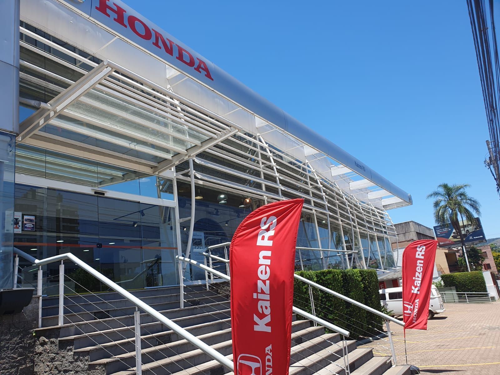 Concessionária Honda com fachada de vidro, bandeiras vermelhas e céu azul.