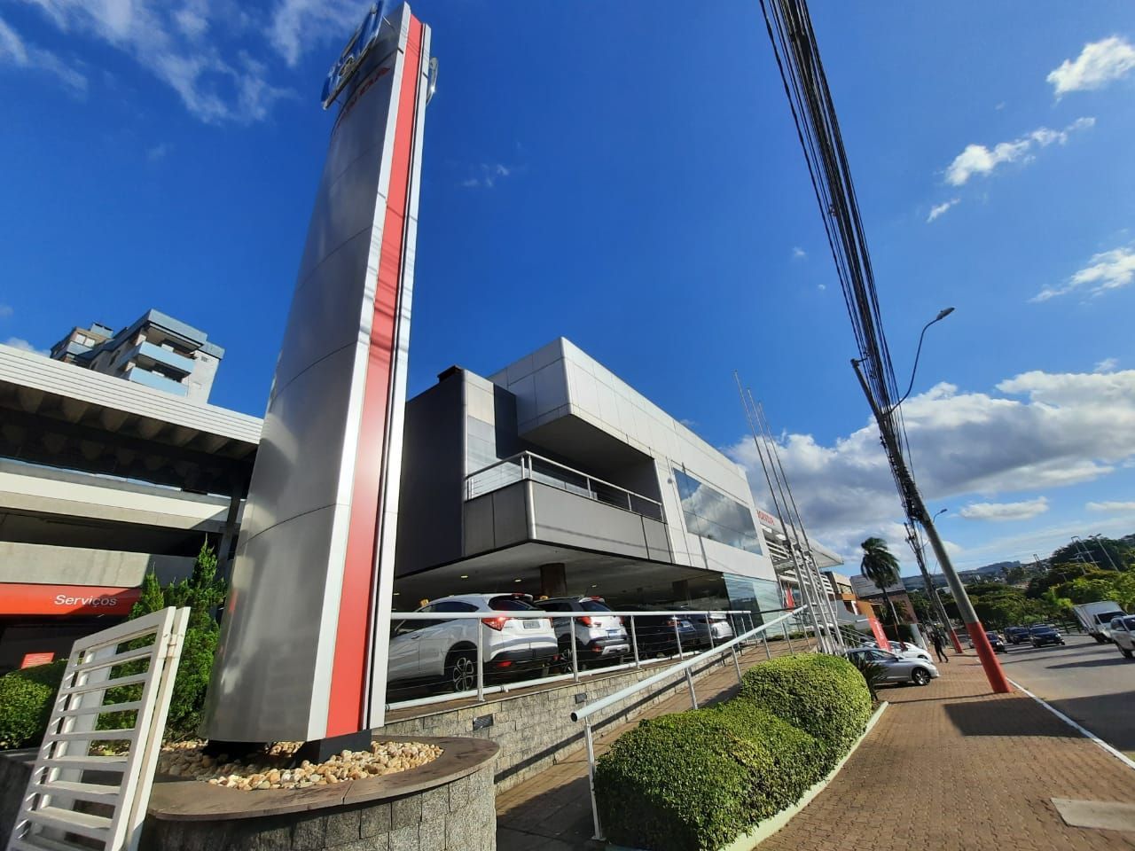 Prédio de concessionária de automóveis com uma placa alta vermelha e prateada, céu azul.