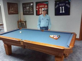 A man is standing next to a pool table in a room with jerseys on the wall.