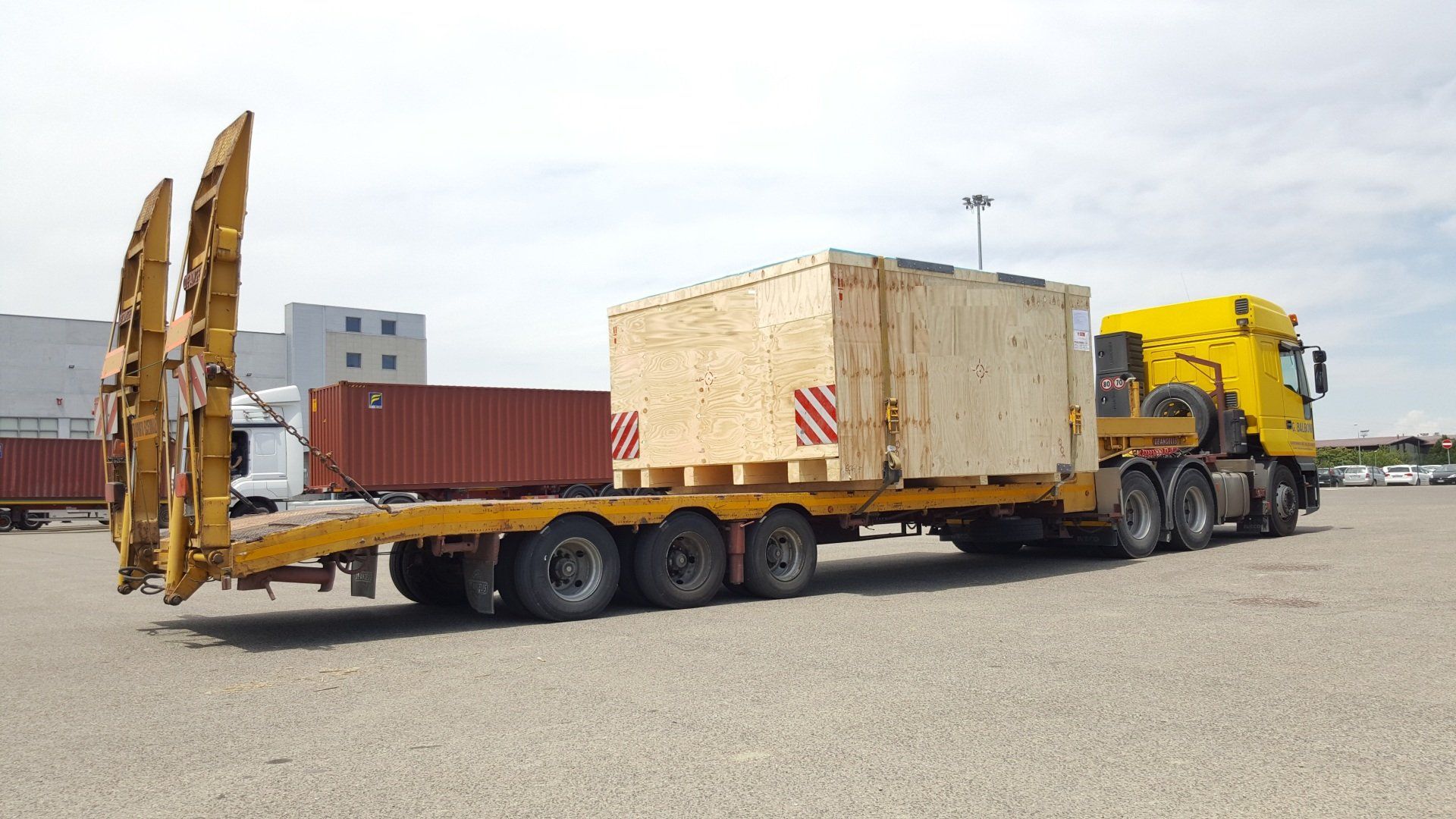 Un camion giallo trasporta una grande cassa di legno e un container su un rimorchio a pianale ribassato in un parcheggio.