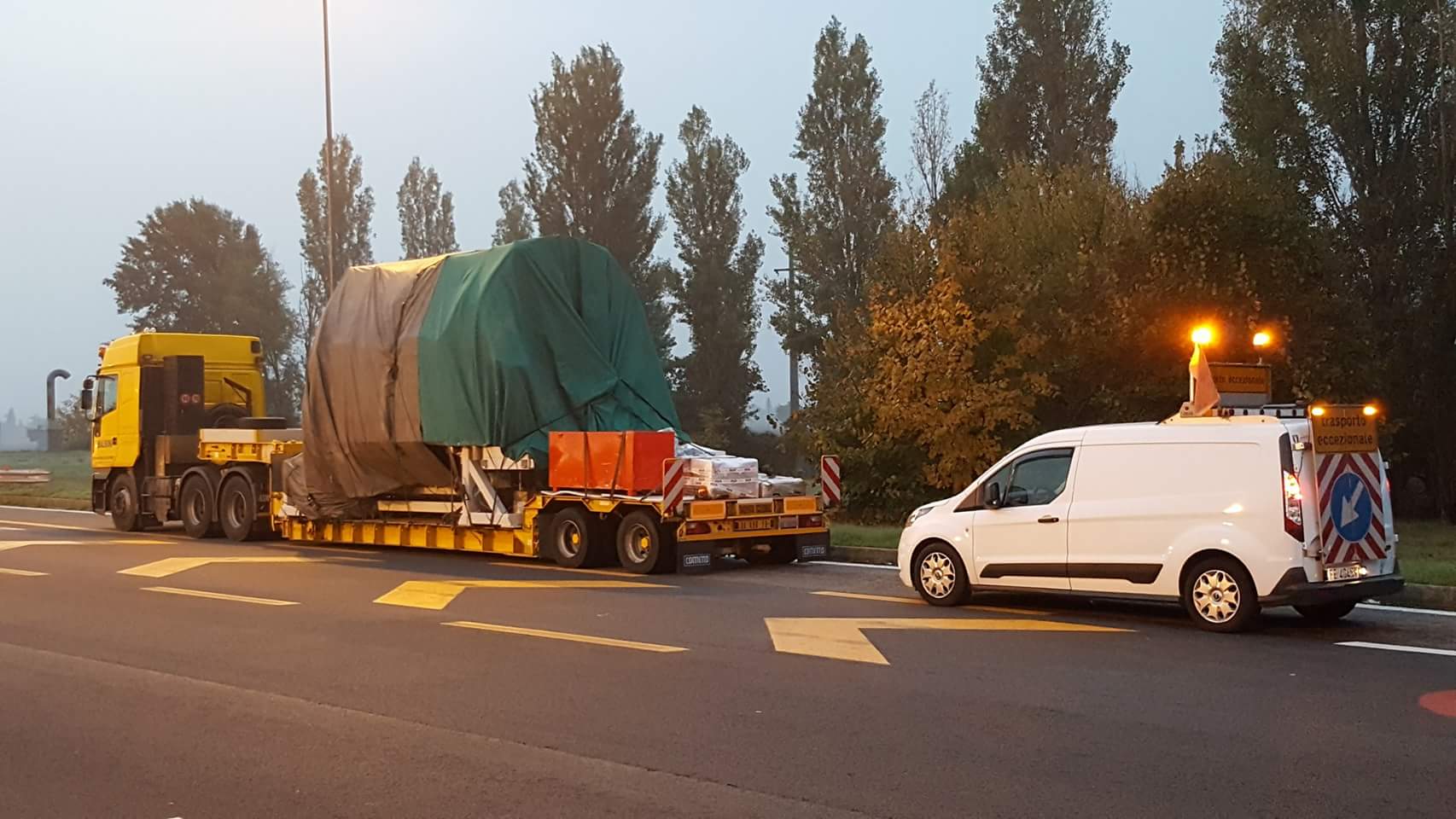 Due camion gialli parcheggiati fuori da un edificio, uno con un rimorchio a pianale ribassato e l'altro con una gru.