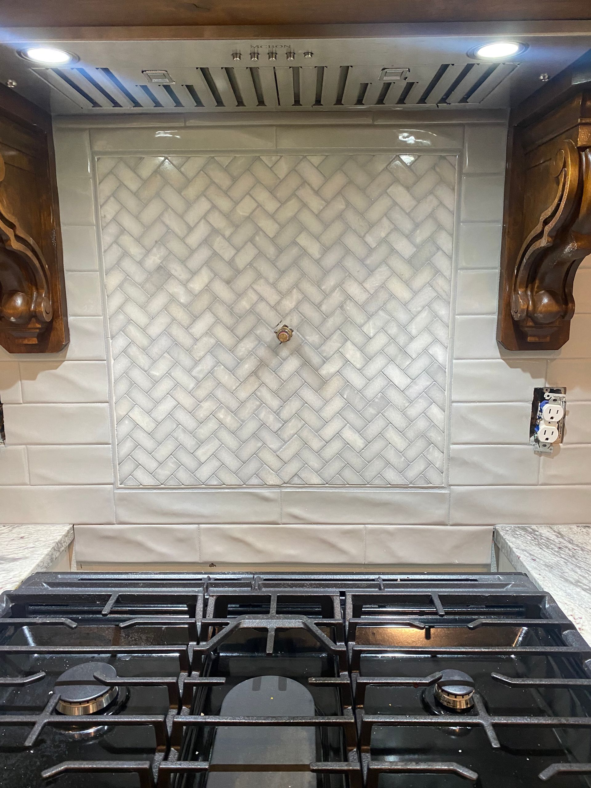 Kitchen backsplash with herringbone pattern, above a gas stovetop. Wooden accents and white and gray tiles.
