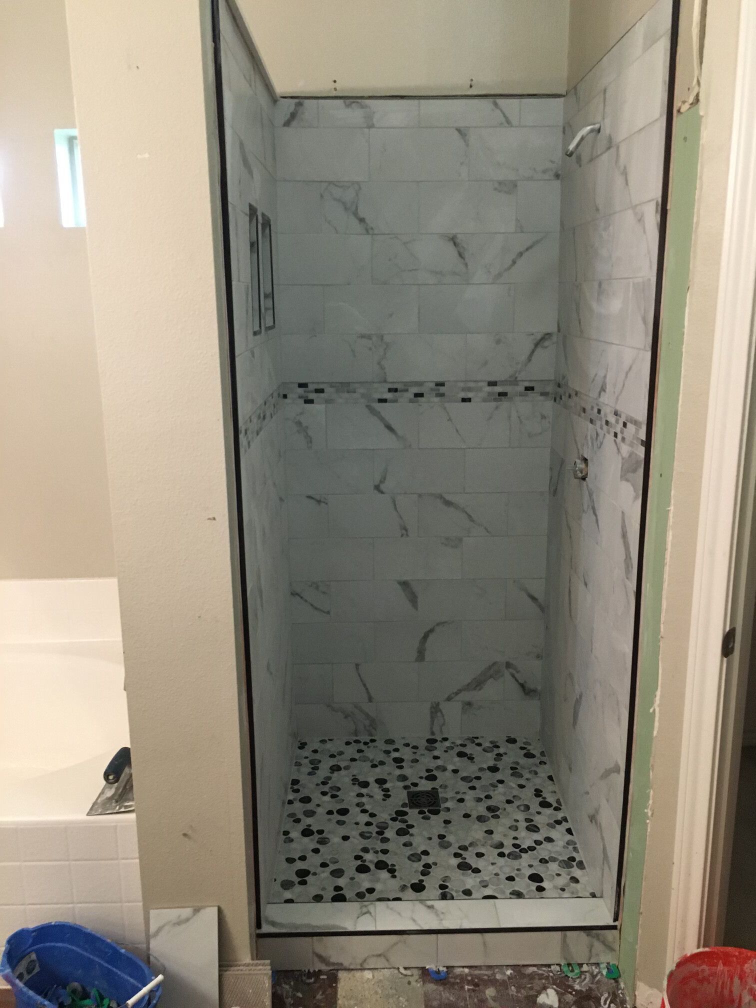 Partially tiled shower stall with white marble-look tiles and black pebble floor.