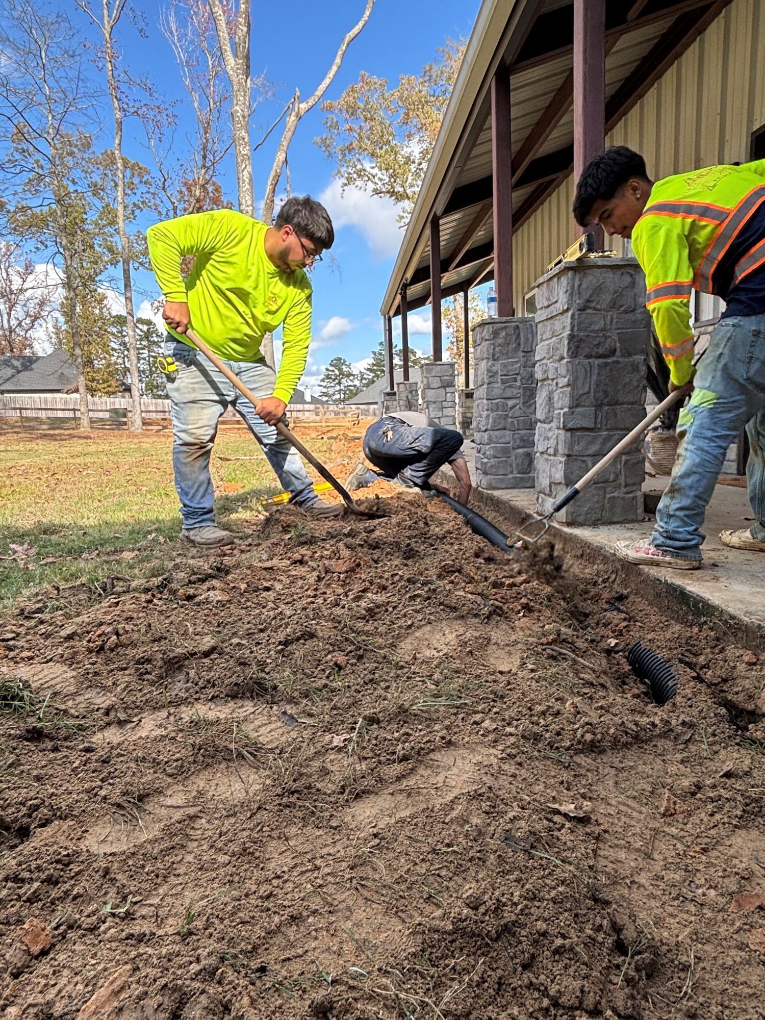 Two workers, wearing neon shirts, are digging a trench near a building with a stone border.