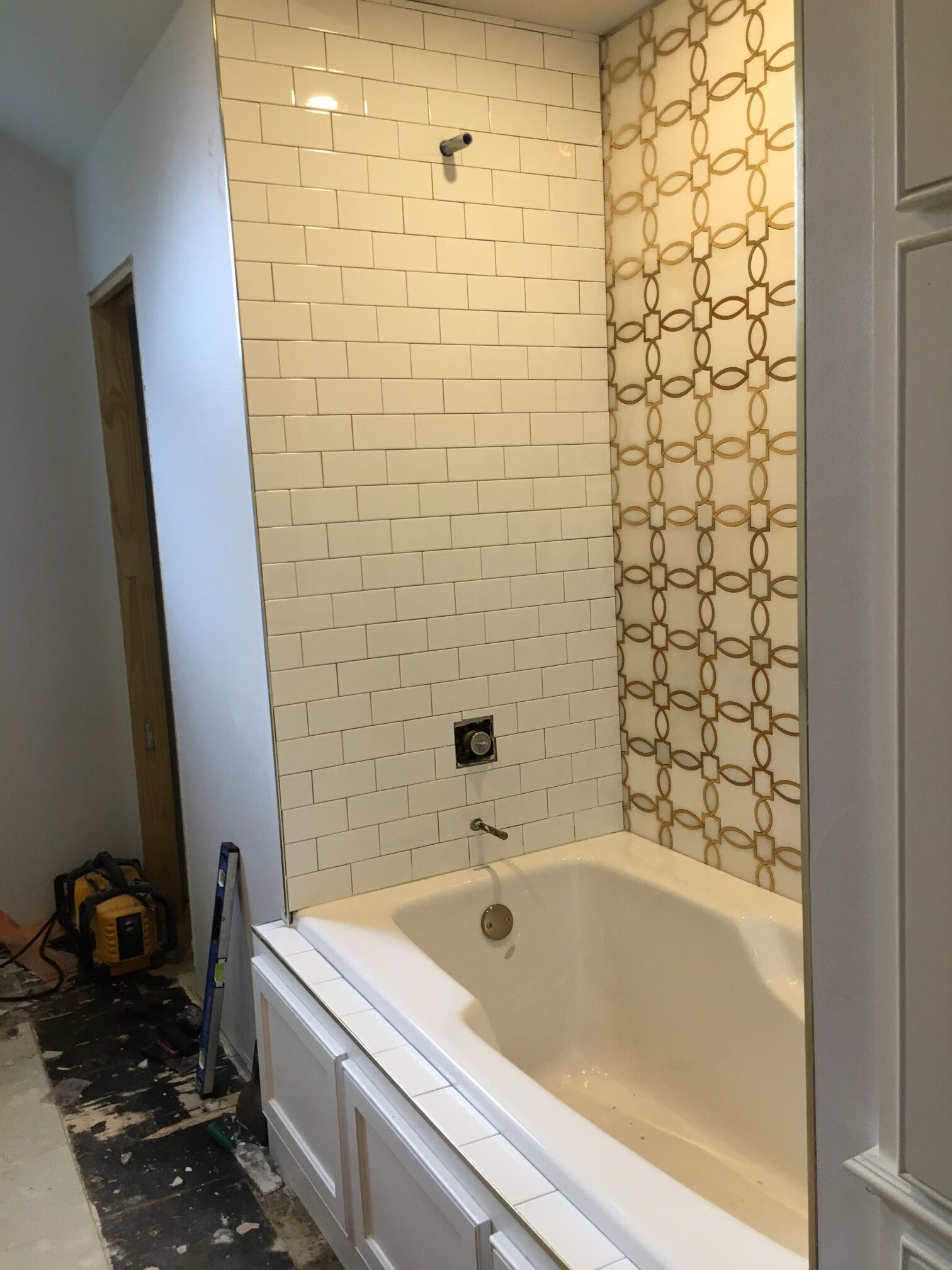Bathroom under construction with white tub, gold-patterned and white tiled walls, and exposed plumbing fixtures.
