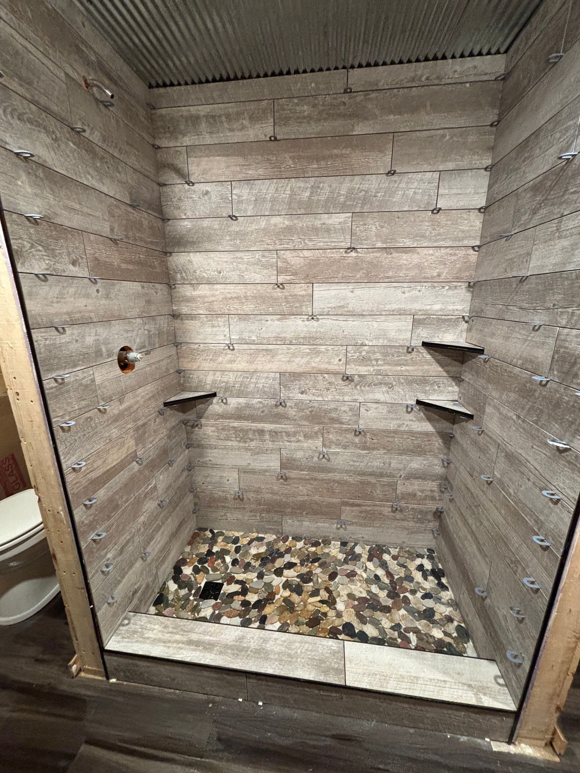 Shower stall with light wood-look tiled walls, pebble floor, and glass enclosure.