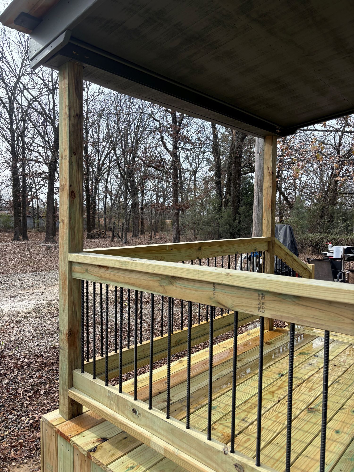 Wooden deck with black railings and covered roof, overlooking a wooded area.