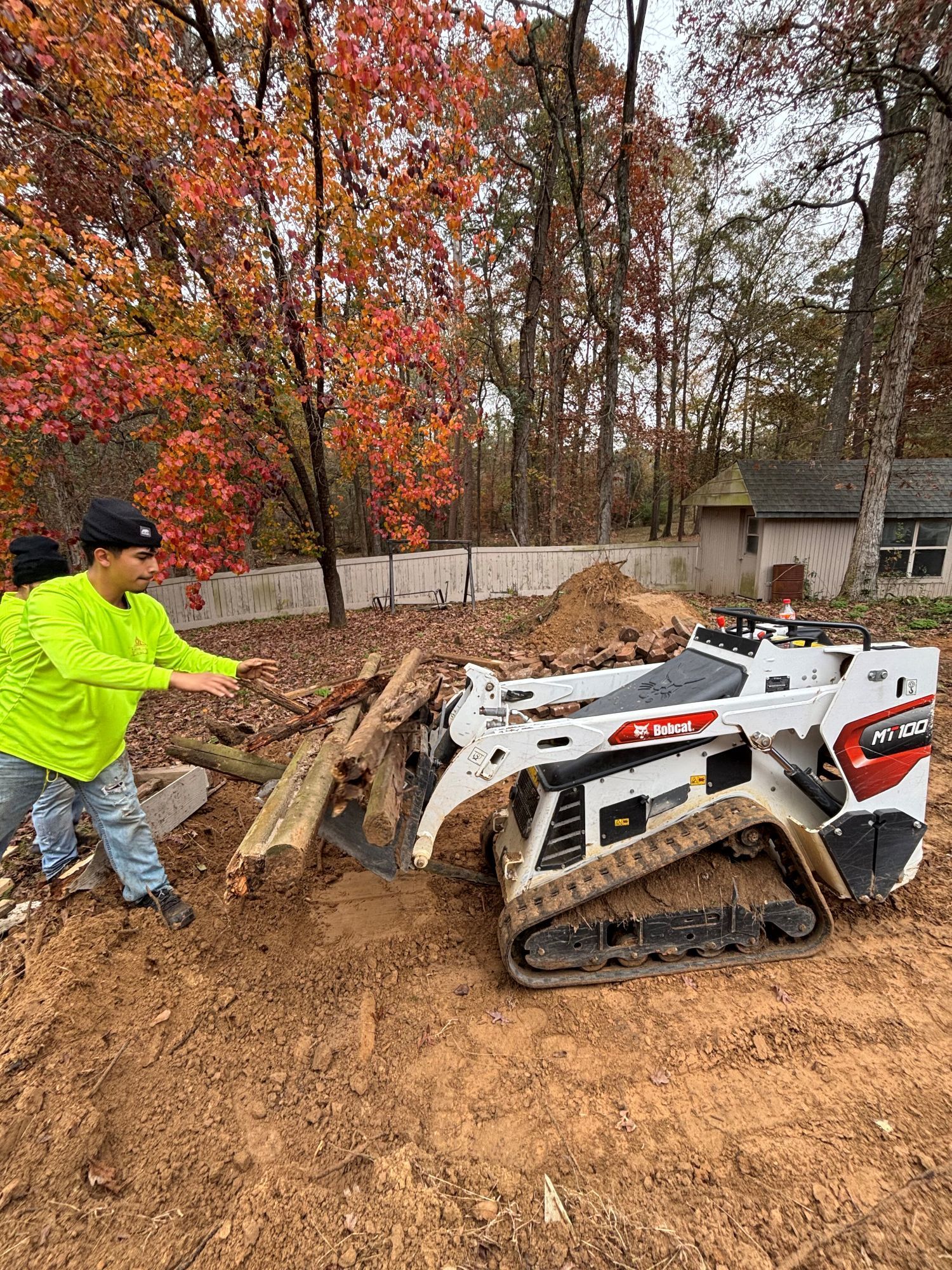 Two people move wood with a Bobcat skid-steer loader outdoors. Fall foliage.