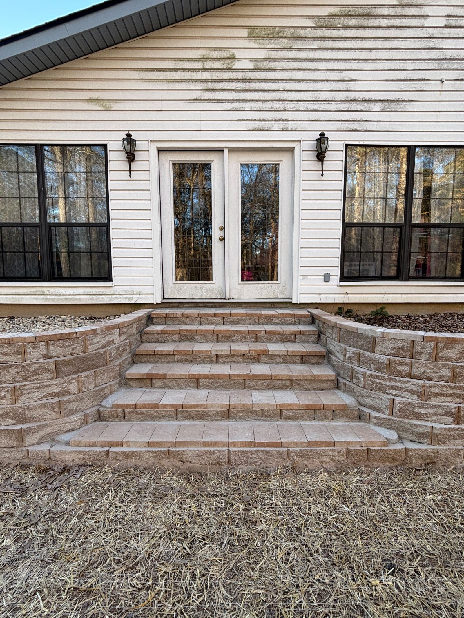 Exterior of a house with steps and double doors; light-colored siding, black window frames.